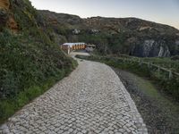 a narrow path that is next to a big cliff side building, near an area with trees and a fence