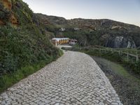 a narrow path that is next to a big cliff side building, near an area with trees and a fence