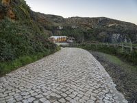 a narrow path that is next to a big cliff side building, near an area with trees and a fence