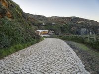 a narrow path that is next to a big cliff side building, near an area with trees and a fence