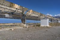 an industrial structure with large solar panels in a desert field near the ocean, along with a bench to watch for fish