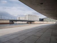 a concrete walkway leading over a bridge and into a building with a sky background and some buildings on both sides