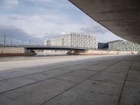 a concrete walkway leading over a bridge and into a building with a sky background and some buildings on both sides