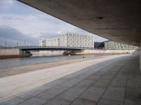 a concrete walkway leading over a bridge and into a building with a sky background and some buildings on both sides