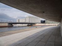 a concrete walkway leading over a bridge and into a building with a sky background and some buildings on both sides