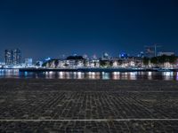 a bench with lights on near the water by the city skyline at night, with a full view across the water