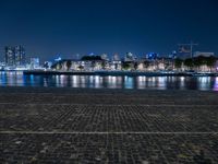 a bench with lights on near the water by the city skyline at night, with a full view across the water