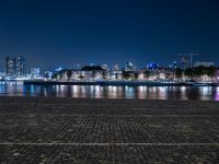 a bench with lights on near the water by the city skyline at night, with a full view across the water