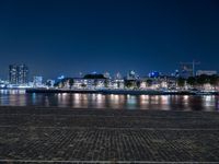 a bench with lights on near the water by the city skyline at night, with a full view across the water