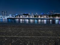 a bench with lights on near the water by the city skyline at night, with a full view across the water