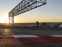 a sun setting over a parking lot near the water with a bridge above it and cars driving on it