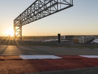 a sun setting over a parking lot near the water with a bridge above it and cars driving on it