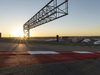 a sun setting over a parking lot near the water with a bridge above it and cars driving on it