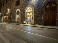 man sitting on a bench at a stone building at night next to a sidewalk that has yellow lines