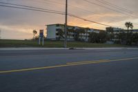 an empty road and a house near some palm trees in the sunset distance with power lines over it