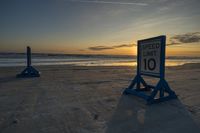 two blue posts sit on the sand at sunset beside the ocean's edge with speed limit sign