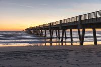 a sandy beach with a pier near by at sunrise with the tide on it and waves coming in