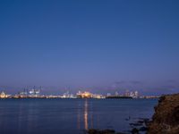 a city is seen across the water at dusk, with buildings in the background on a rocky coast