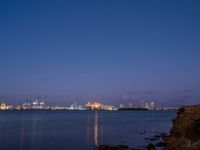 a city is seen across the water at dusk, with buildings in the background on a rocky coast