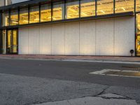 a street with an asphalt lot next to a concrete building and glass windows with a light on each floor