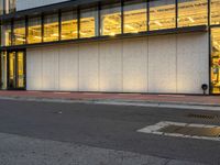 a street with an asphalt lot next to a concrete building and glass windows with a light on each floor