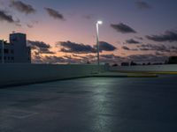 a parking lot with a building in the background and street lamps lite up at dusk