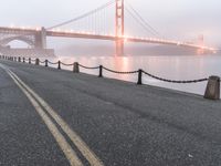 an empty street beside the water and a bridge with fog covering it from sun and fog