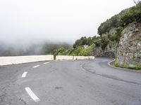 curved road in the middle of a foggy hillside covered with rock, surrounded by fog, and green trees