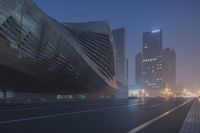 cars drive down a foggy street with tall buildings behind it at night in front of tall towers