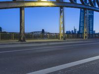 the underside of a steel bridge at dusk, with city buildings in the background at night