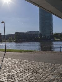 a city scene with a large tower and a lake in the foreground and a person sitting on a bench with an umbrella near it