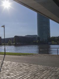 a city scene with a large tower and a lake in the foreground and a person sitting on a bench with an umbrella near it