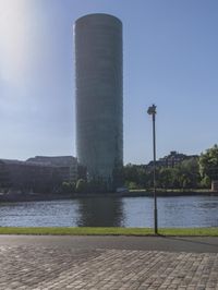 a city scene with a large tower and a lake in the foreground and a person sitting on a bench with an umbrella near it