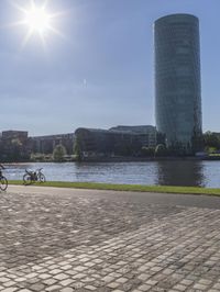 a city scene with a large tower and a lake in the foreground and a person sitting on a bench with an umbrella near it