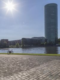 a city scene with a large tower and a lake in the foreground and a person sitting on a bench with an umbrella near it