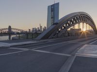 the sun is setting over the roadway in front of the bridge crossing river bank with downtown skyline behind