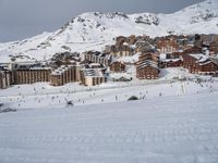 people snowboarding on a very big snowy mountain slope with buildings nearby and many tracks in the snow