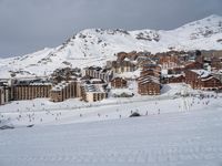 people snowboarding on a very big snowy mountain slope with buildings nearby and many tracks in the snow