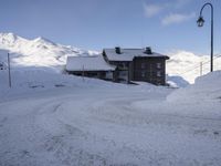 an empty road covered in snow on the slopes of a mountain top, with houses and mountains behind it