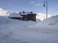 an empty road covered in snow on the slopes of a mountain top, with houses and mountains behind it