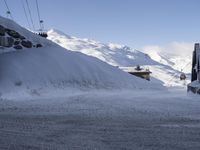 a snowy road in front of the snow - capped mountains with ski lift and lodge nearby