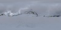 the skier is standing in the snow under a large mountain with trees on it and clouds rolling in