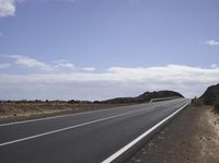 a highway in the desert with a hill in the background of blue skies and clouds
