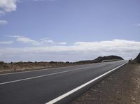 a highway in the desert with a hill in the background of blue skies and clouds