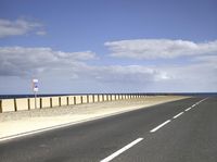 a road and an ocean in a blue sky with white clouds, next to a wall