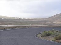 an empty curved road leading to the village below it with some hills in the background