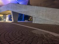 a paved path at night leading to an illuminated building with colorful lights and pillars in a lit city