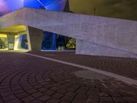 a paved path at night leading to an illuminated building with colorful lights and pillars in a lit city