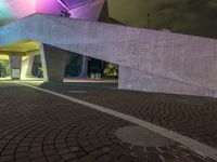 a paved path at night leading to an illuminated building with colorful lights and pillars in a lit city