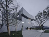 some trees and some green grass bushes and lights in the evening sky at a concrete building with large windows